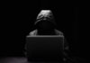 A hooded figure sitting in front of a laptop in a dark setting