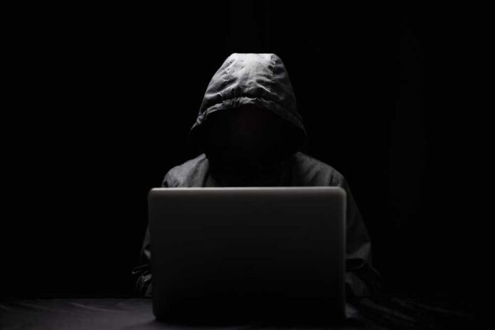 A hooded figure sitting in front of a laptop in a dark setting