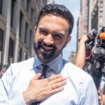 Shelter Standoff: Progressive Mayor Faces Backlash A smiling man in a light blue shirt gestures with his hand in an outdoor urban setting
