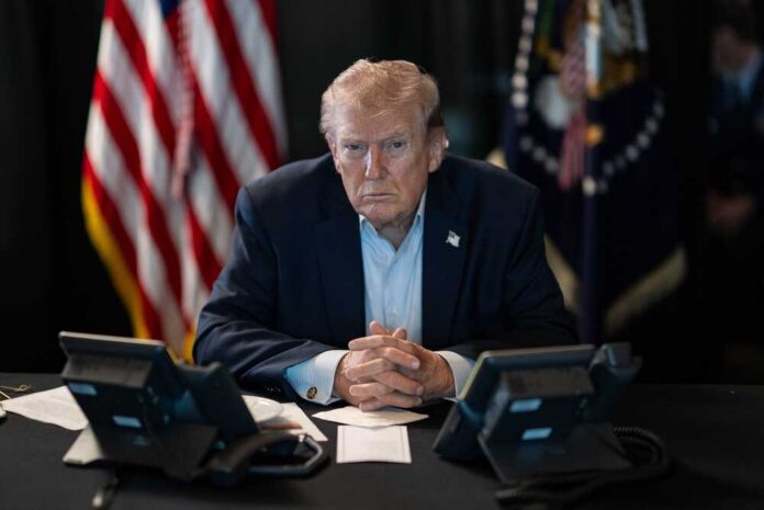 A serious-looking man seated at a table with phones and an American flag in the background