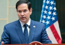 A man in a blue suit speaking into a microphone with an American flag in the background