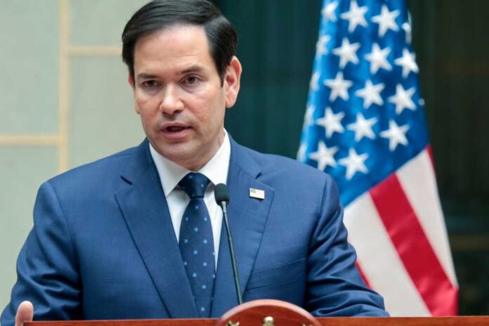 A man in a blue suit speaking into a microphone with an American flag in the background