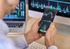 Person checking stock market graphs on phone and monitors.