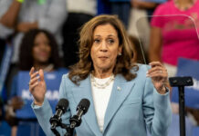 Woman in light blue blazer speaking at podium with microphones