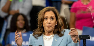 Woman in light blue blazer speaking at podium with microphones