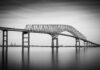 Baltimore’s Bridge Collapse: Hidden Truths Emerge A large bridge spanning over a calm body of water in black and white
