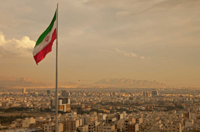 Iran,Flag,Waving,In,The,Wind,Above,Skyline,Of,Tehran Iranian flag waving over a city skyline with mountains in the background