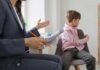 A child sitting on a chair while a professional gestures with a notepad and pencil