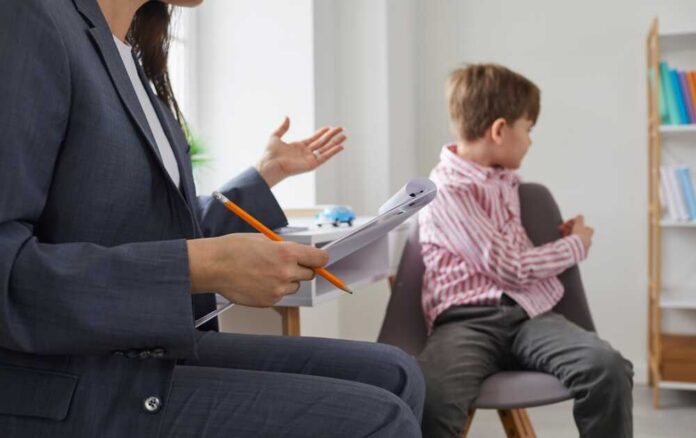 shutterstock_2366266423.jpg A child sitting on a chair while a professional gestures with a notepad and pencil