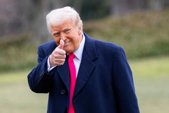 A man in a dark coat and pink tie giving a thumbs up outdoors