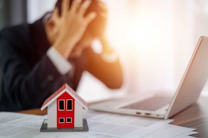Businessman,Headache,Bad,Economy,And,Inflation,Crisis,Leads,To,Rise A businessman with his head in his hands, stressed over paperwork and a model house on the table