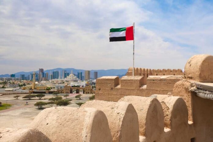 United,Arab,Emirates,Flag,Flying,Above,Fujairah,Fort,With,City UAE flag waving atop a historic fort with a city skyline in the background