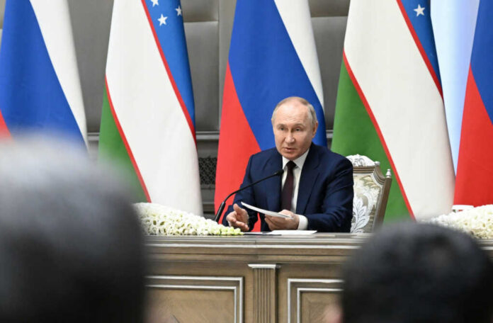 man in suit speaking at a conference table with microphones and flags behind