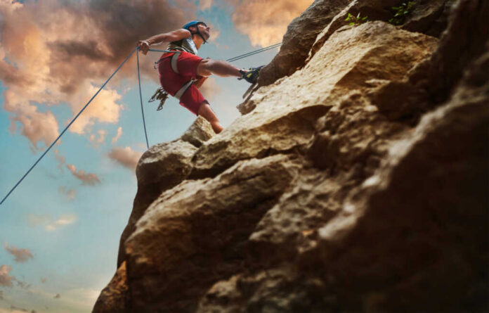 climber-man-in-protective-helmet-abseiling-from-cliff2165035305jpg Person rock climbing with ropes against a cloudy sky