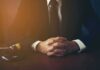 person in suit with hands clasped at a desk beside a gavel
