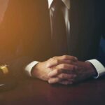 person in suit with hands clasped at a desk beside a gavel