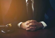 Probe Erupts After ‘Full Accountability’ Boast person in suit with hands clasped at a desk beside a gavel