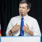 Power Grab? Buttigieg Targets Rural Clout man in white shirt and blue tie speaking at a podium