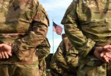 Soldiers in camouflage stand with hands behind backs facing a U.S. flag