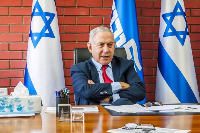 man in suit seated at a desk with Israeli flags behind him