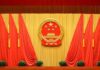 China’s Espionage Charge – Beijing Grabs American Chinese national emblem on red-draped stage backdrop
