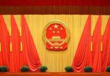 China’s Espionage Charge – Beijing Grabs American Chinese national emblem on red-draped stage backdrop