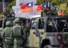 Soldiers beside camouflaged military vehicles with mounted guns and flags
