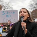 AOC’s Brand Backfires – Branded Toxic In Swing Seats woman speaking into a microphone at an outdoor protest