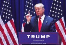 man in suit speaking at podium with multiple American flags