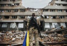 Soldier with rifle stands before a destroyed building holding a blue and yellow flag