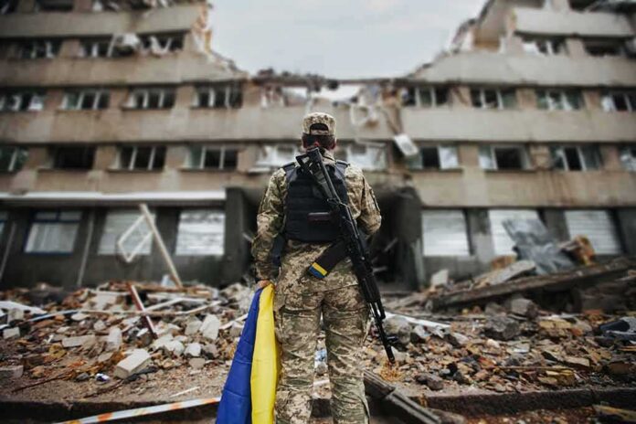 Soldier with rifle stands before a destroyed building holding a blue and yellow flag