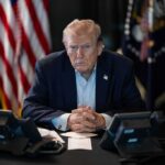 Late-Night Bombshell Targets President Donald Trump seated at a table between two desk phones