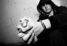 Repeat Gun Kids – Juvenile Loop Exposes System Gaps A young person in a hooded sweatshirt sitting against a wall with hands clasped and handcuffs visible