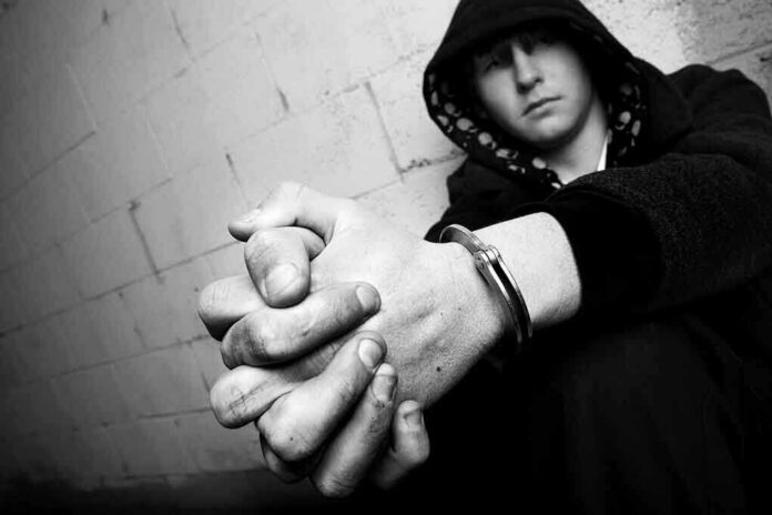 shutterstock_9474589 A young person in a hooded sweatshirt sitting against a wall with hands clasped and handcuffs visible